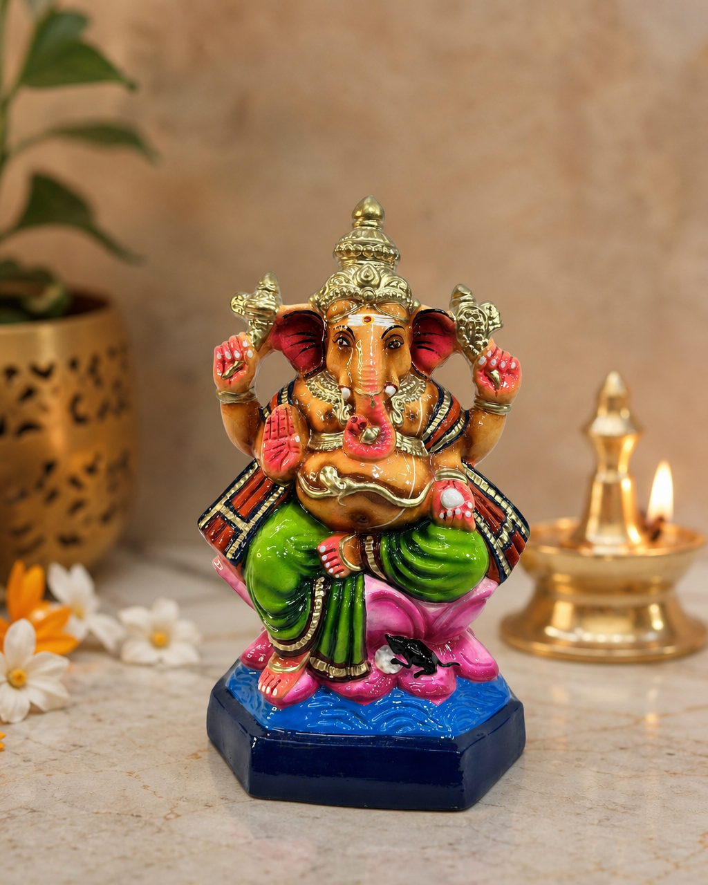 Colorful Vinayagar figurine seated on a decorative base, with vibrant green and pink attire, surrounded by flowers and a diya