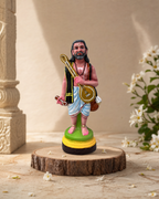 Colorful Kanagadaasar figurine displayed on a wooden base with floral accents.