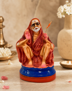 Colorful Kanji Periyavar statue with a red robe and stick, surrounded by festive decor.