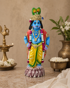 Colorful statue of Vitobha Karnan with blue skin, gold decorations, and a peacock headdress, placed indoors with traditional decor.